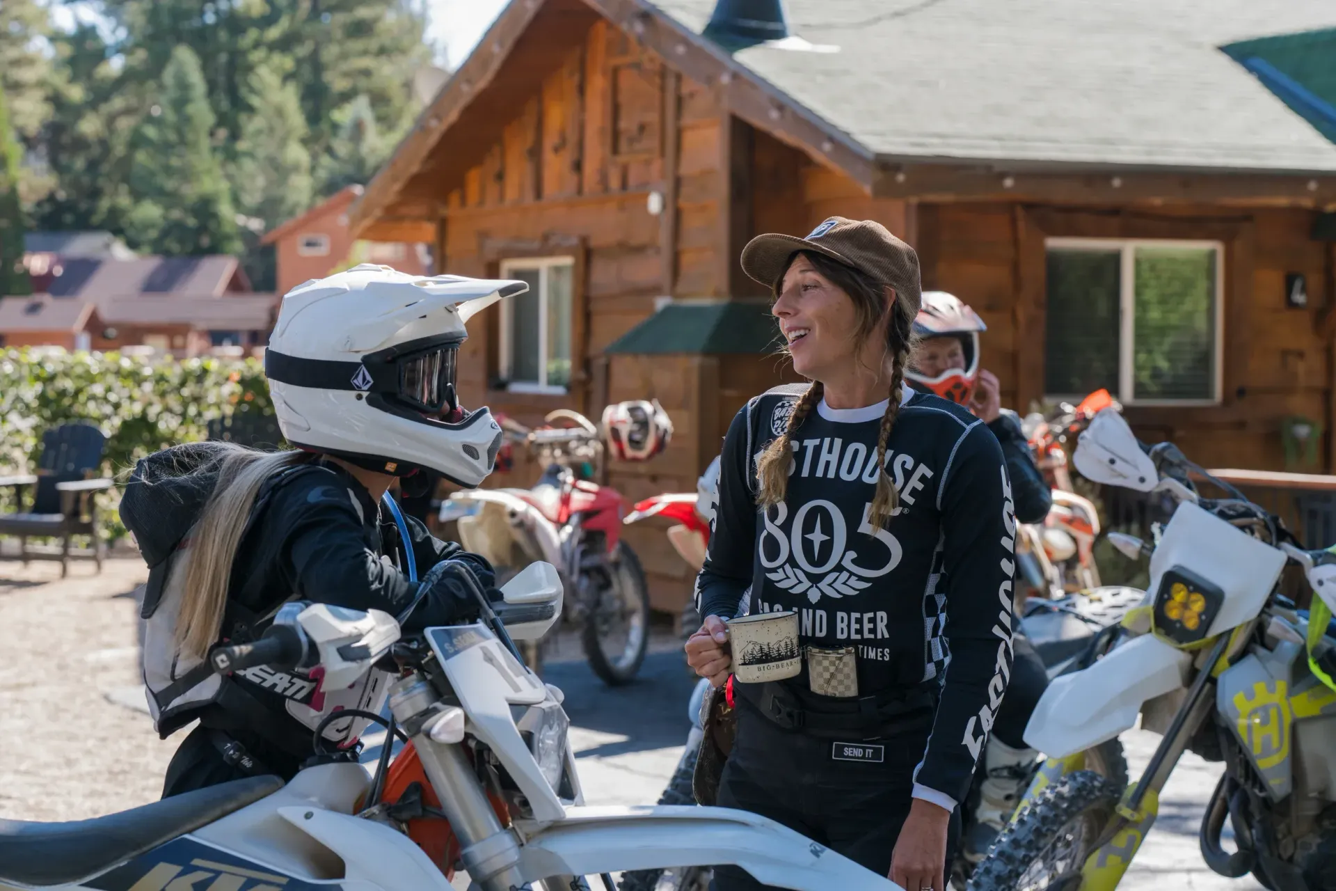 Two dirt bikers conversing near a cabin, motorcycles nearby. One in helmet, the other holding a mug, both smiling.