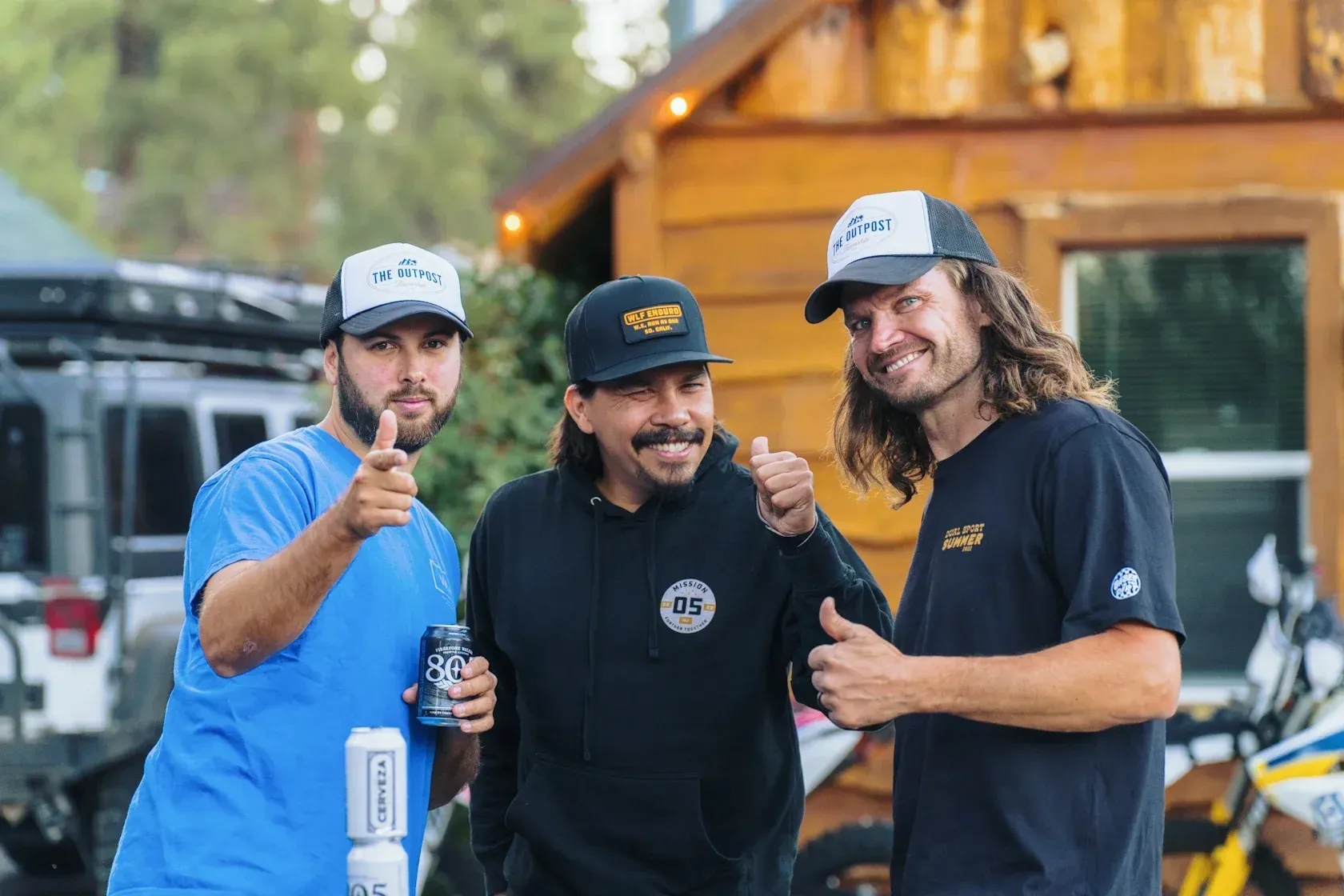 Three men outdoors, giving thumbs up, smiling. One holds a beverage, wearing hats and casual shirts, near a cabin.