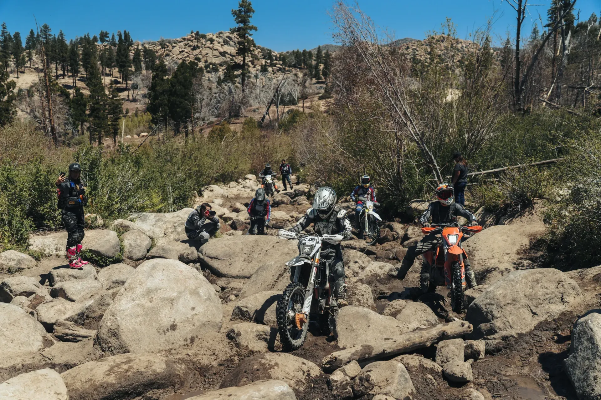Dirt bikes navigating a rocky, muddy trail in a mountainous area under a sunny sky.