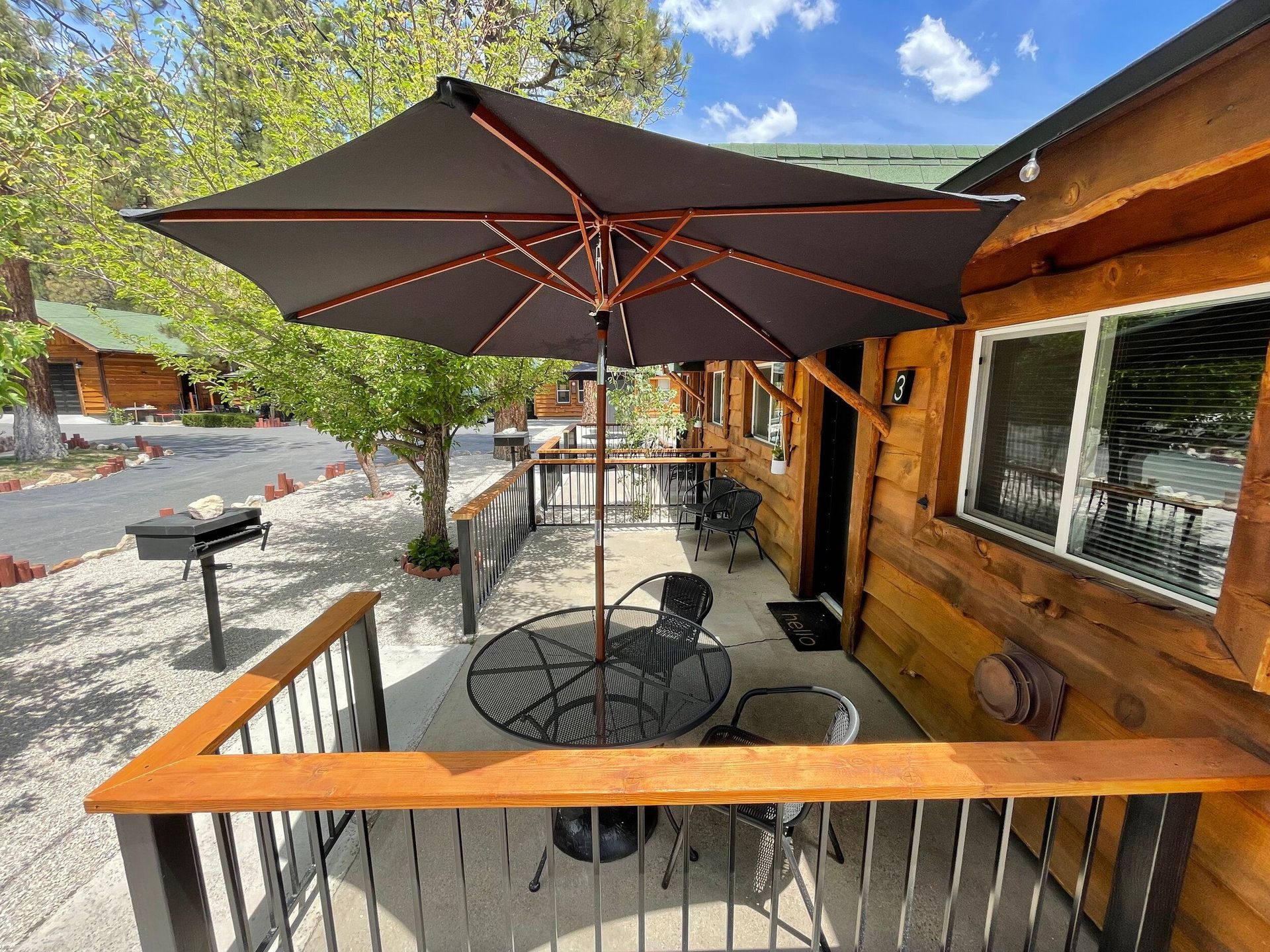 Wooden cabin porch with umbrella, table, and chairs. Grill visible. Sunny outdoor setting.