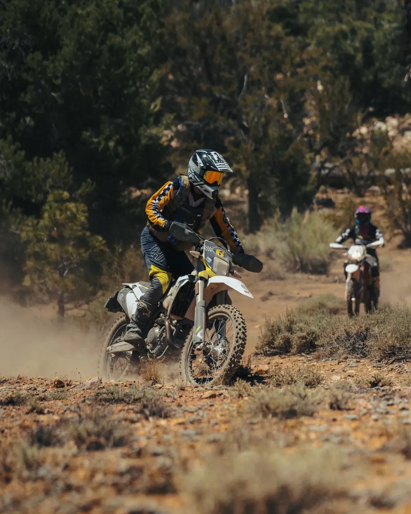 Motorcyclist riding a dirt bike on a dusty trail, another bike in the background, sunny landscape.