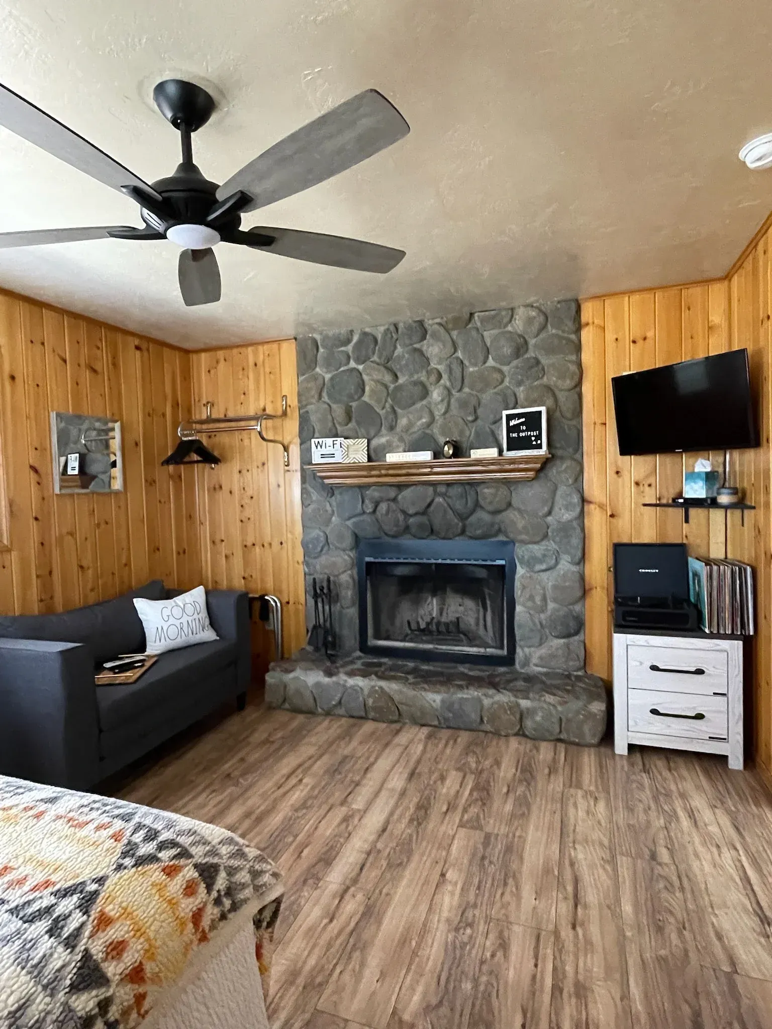 Cozy cabin interior with stone fireplace, wood paneling, dark gray sofa, and ceiling fan.