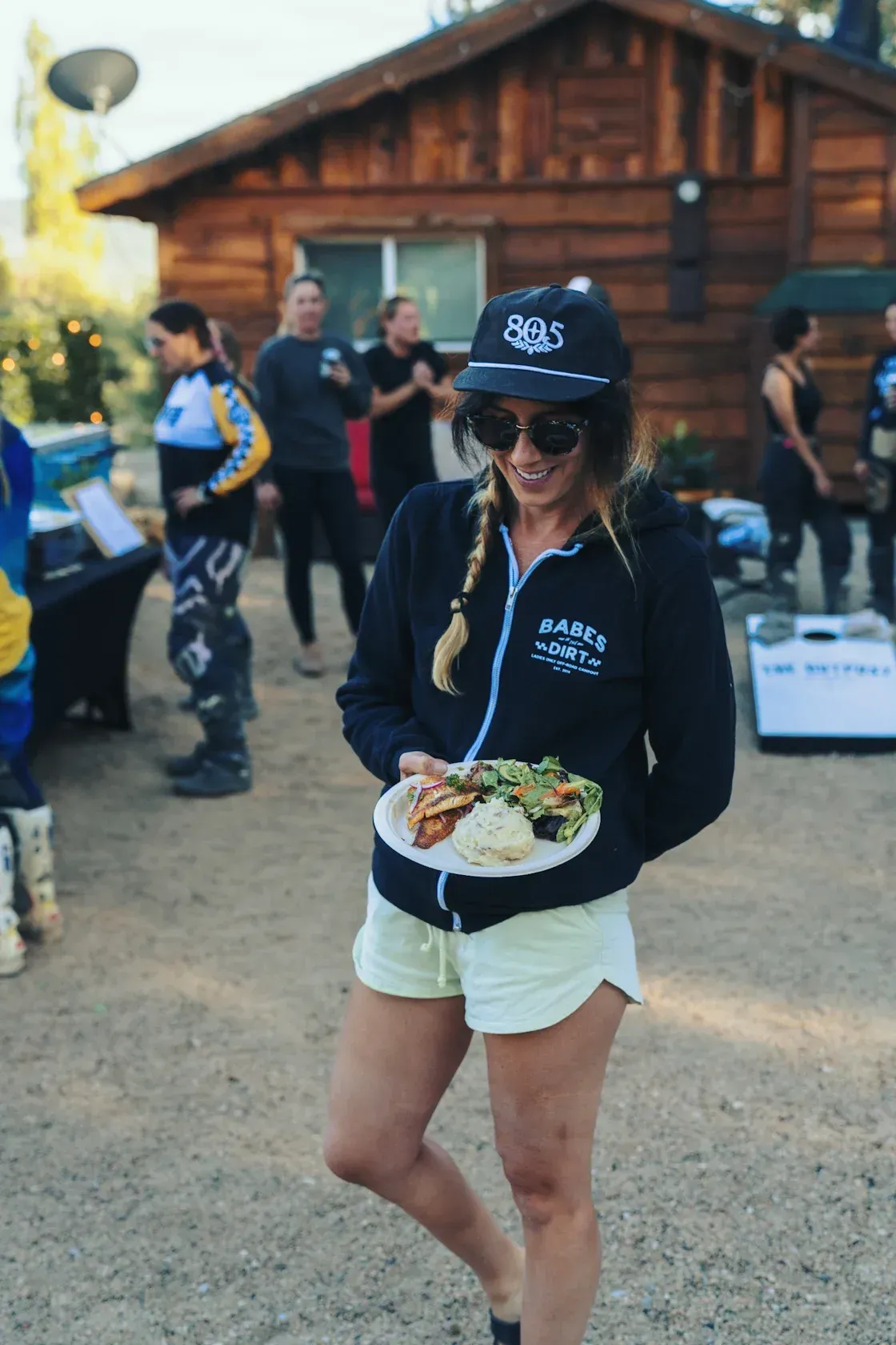 Woman in shorts and hat holding plate of food outside a cabin.