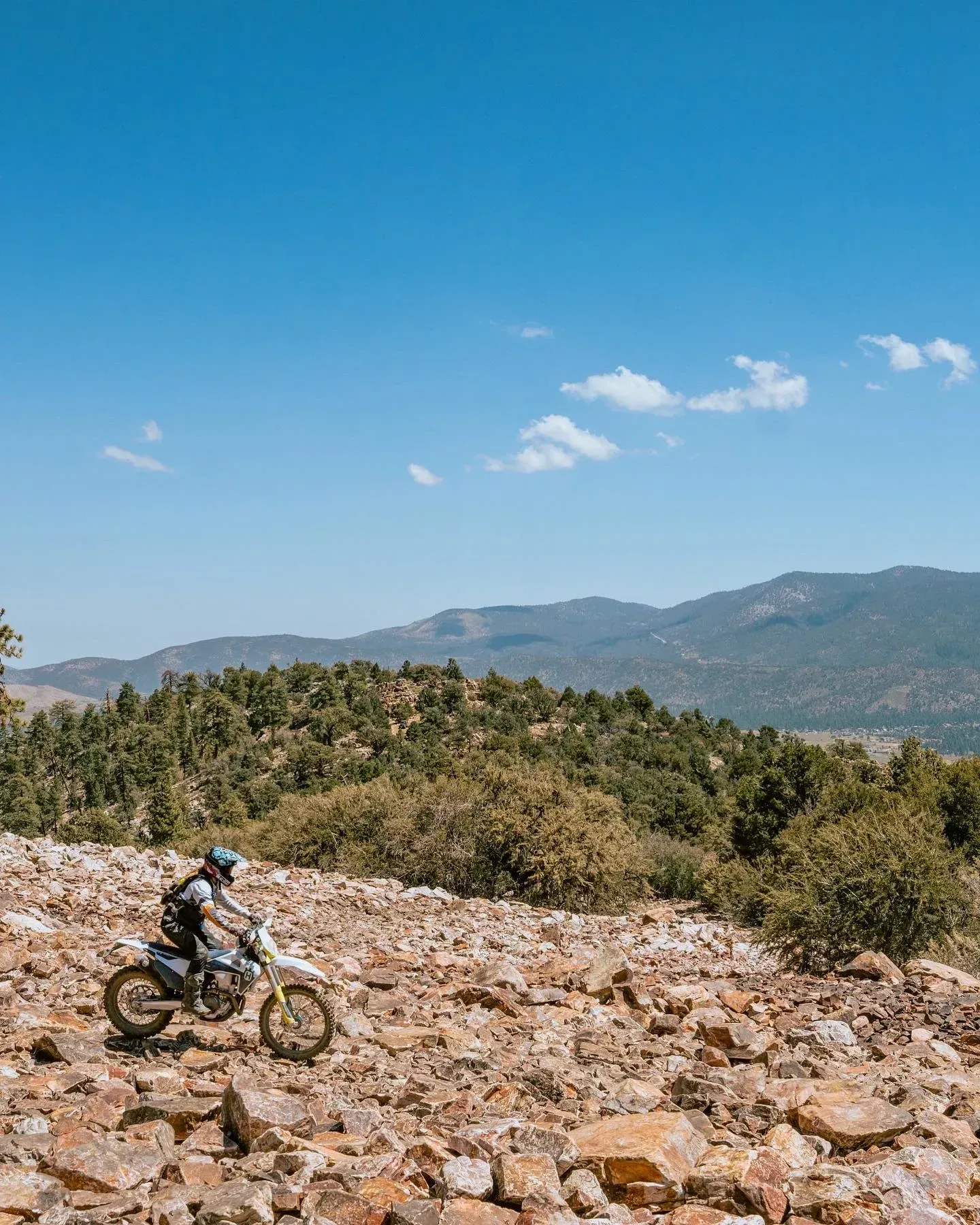 Motorcyclist riding on a rocky dirt path through a mountain landscape under a bright blue sky.