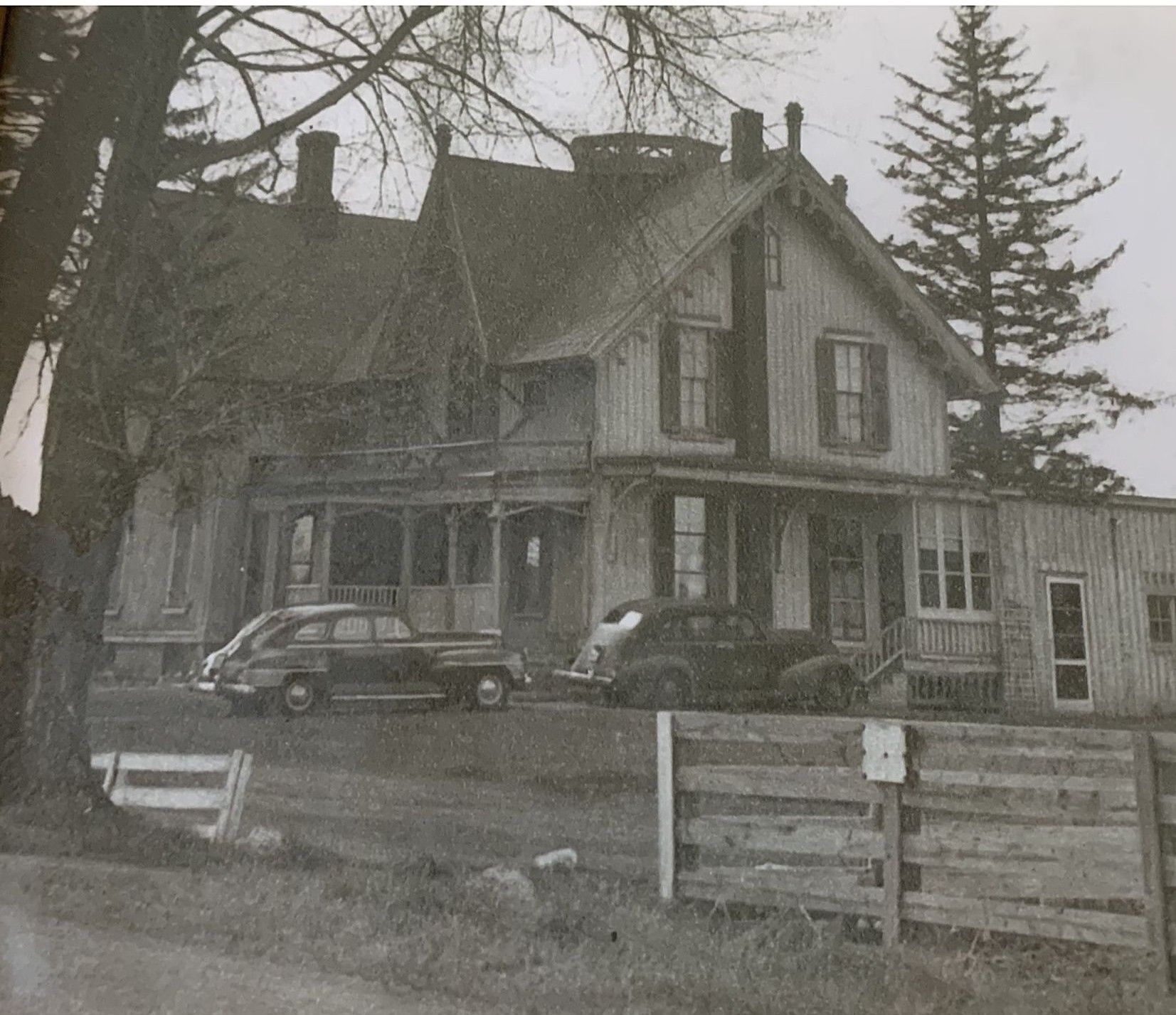 A historic two-story farmhouse with a porch and two vintage cars parked in front, set against trees and a wooden fence.