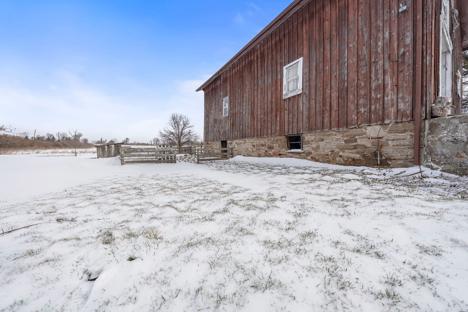 A weathered red wooden barn with a stone foundation stands on a snow-covered field under a bright blue sky.