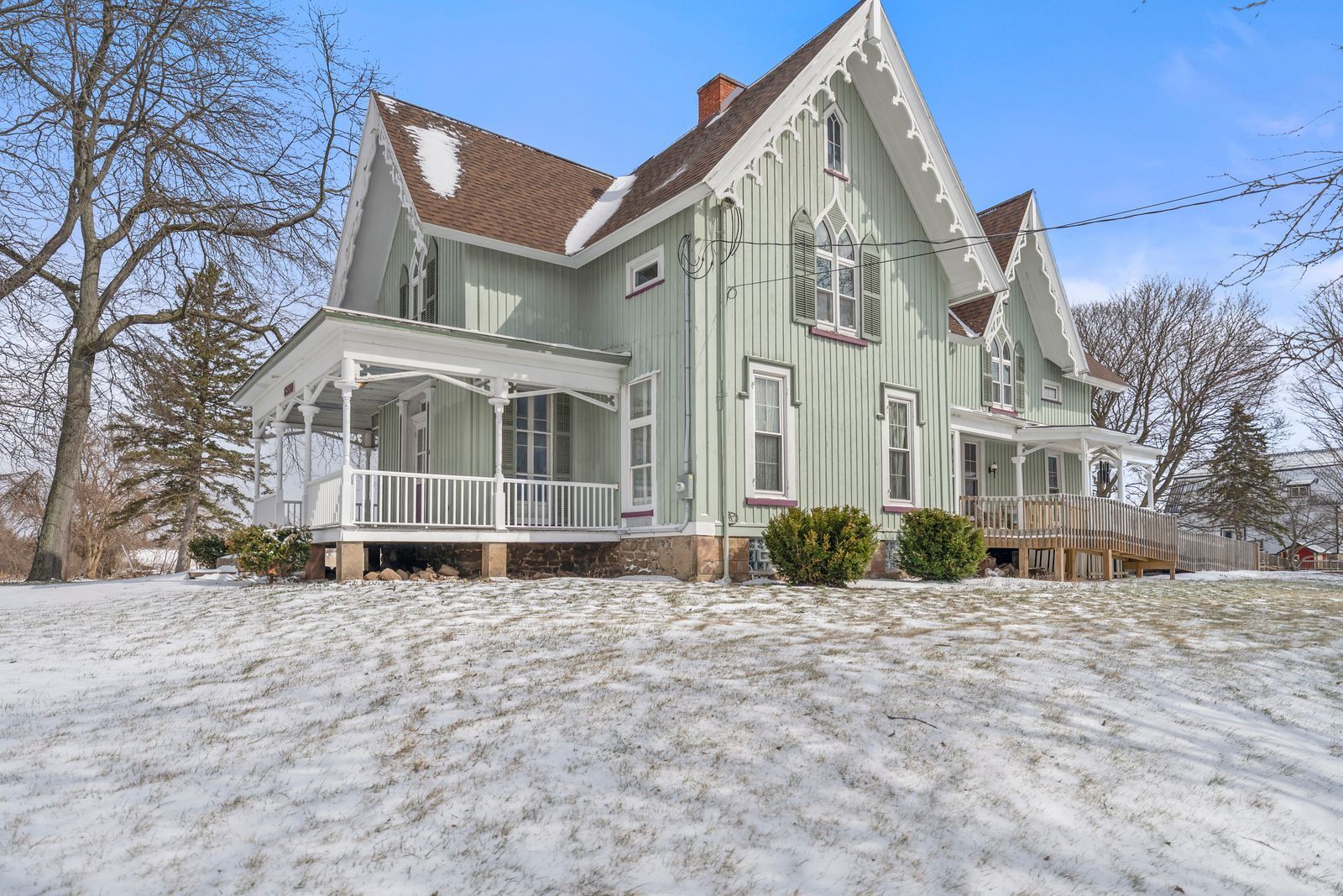 A light green, two-story Gothic Revival house with white trim, a wrap-around porch, and a snowy yard under a blue sky.