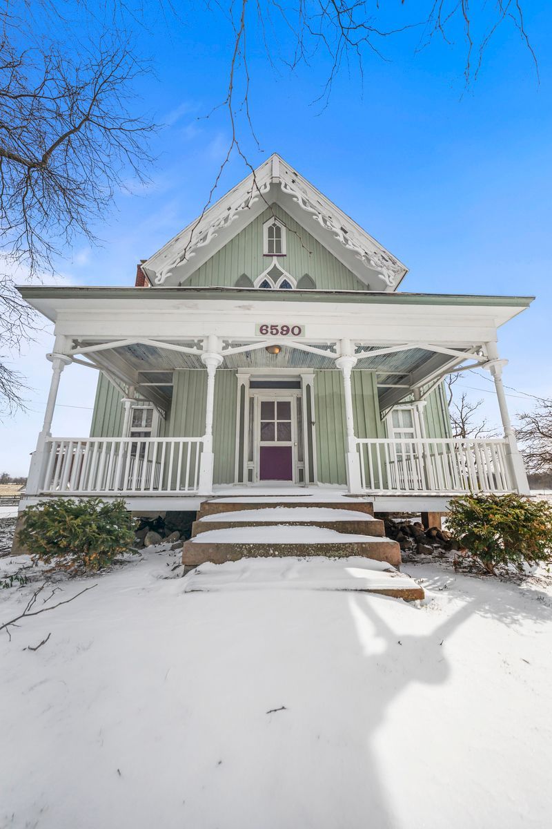 A light green, two-story Gothic Revival house with a white porch and decorative trim, set against snow under a blue sky.