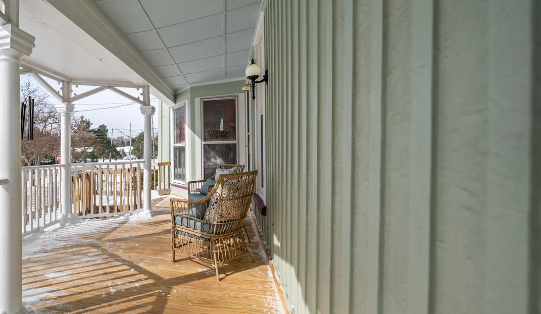 A covered porch with light green vertical siding, a white railing, and a pair of wicker chairs on a snow-dusted floor.