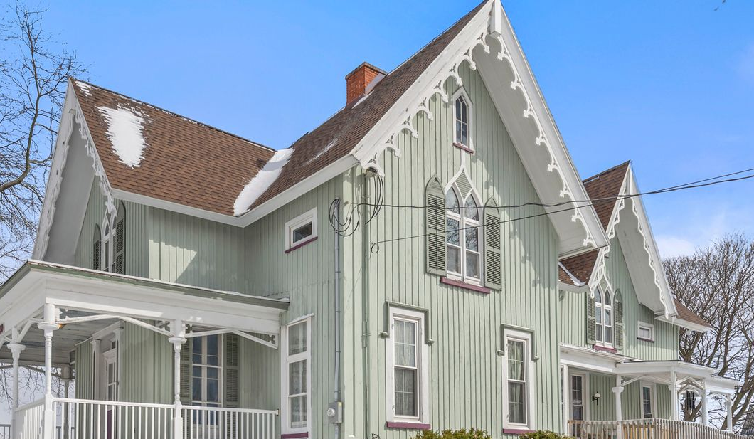 A light green, two-story Victorian-style house with steep gables, ornate trim, and a porch under a clear blue sky.