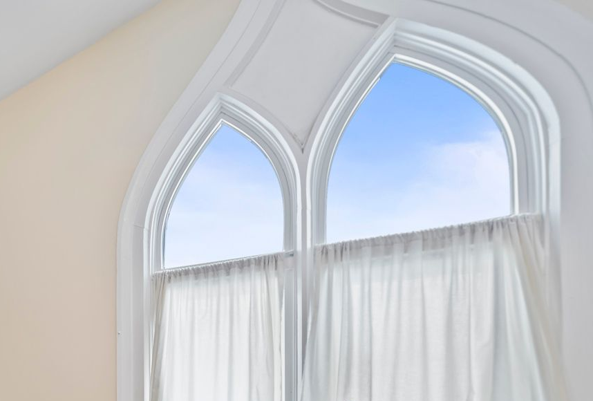 A double arched window with white trim and sheer white curtains looking out onto a bright blue, clear sky.