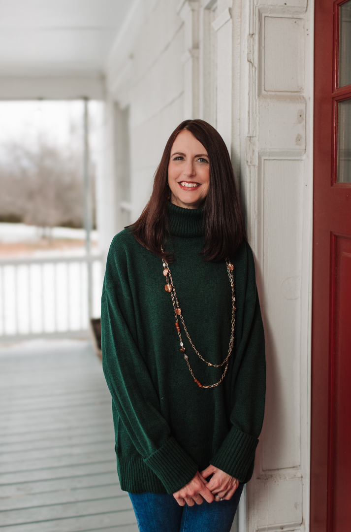A smiling person wearing a dark green turtleneck and layered necklace leans against a white porch post next to a red door.