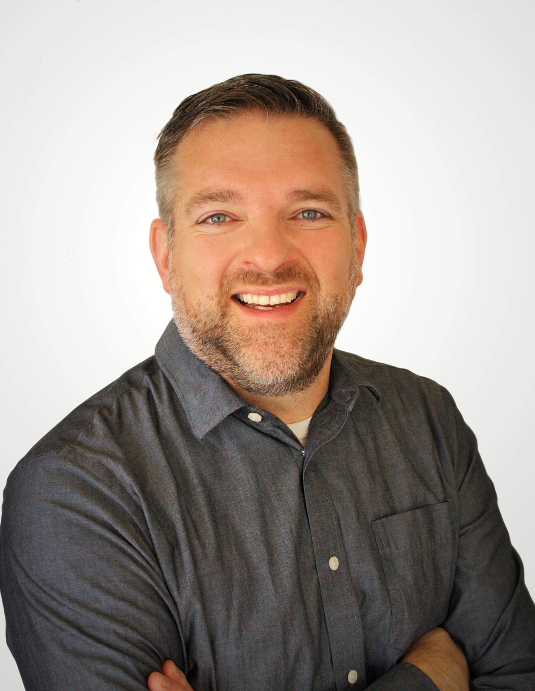 A person with short hair and a beard smiles warmly, wearing a dark button-down shirt with arms crossed against a white wall.