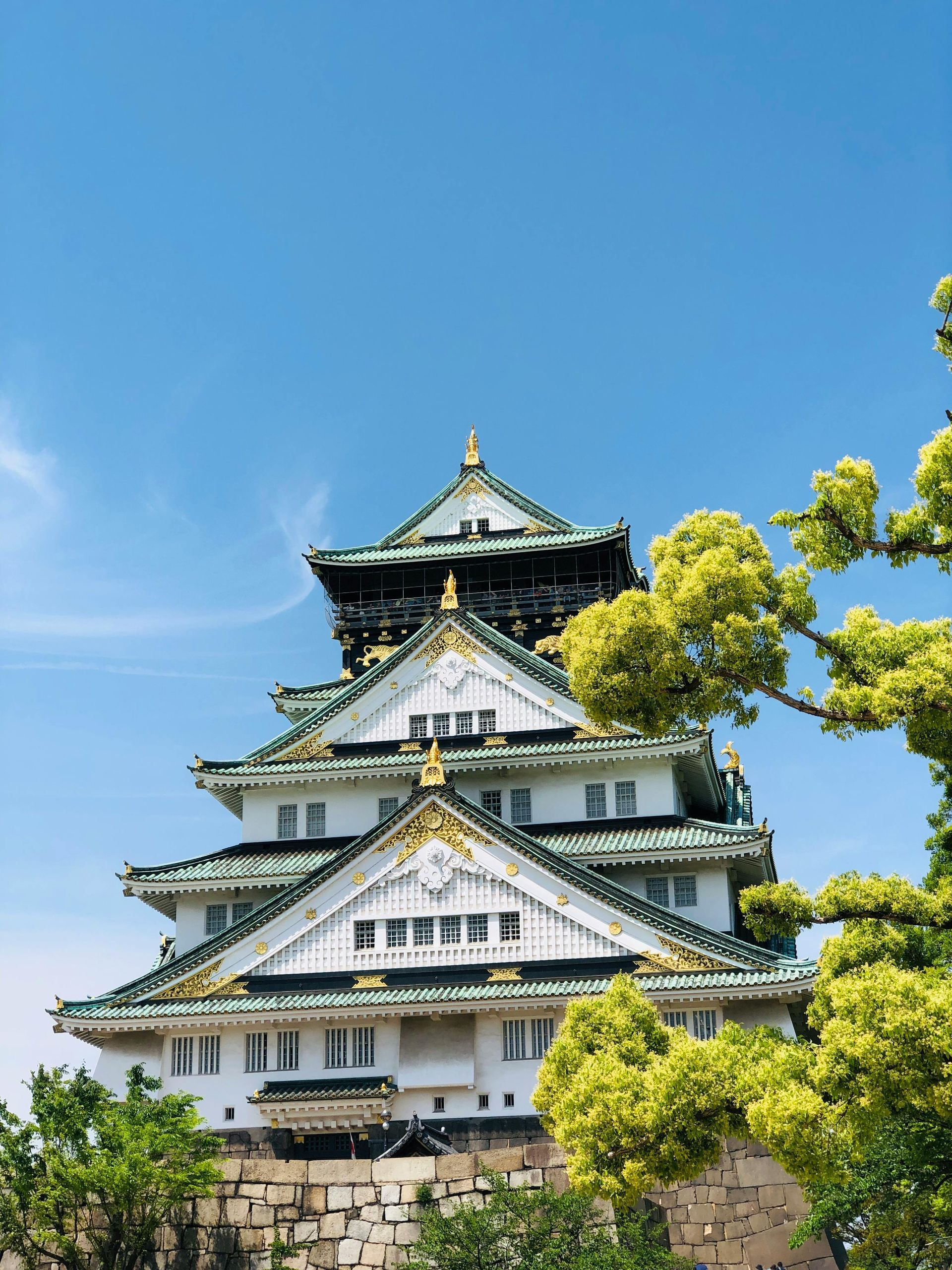Het iconische kasteel van Osaka in Japan, met zijn witte muren, gouden accenten en groene daken onder een helderblauwe hemel.
