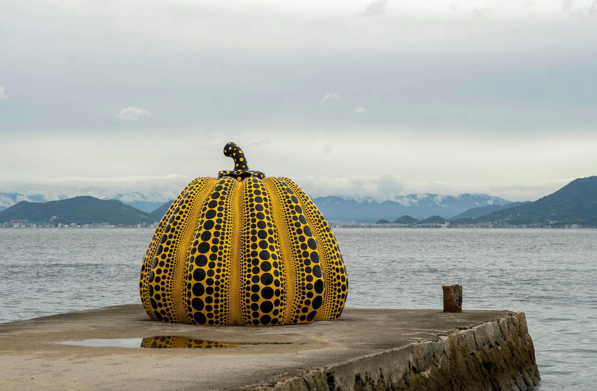 Een grote, gele pompoensculptuur met zwarte vlekken staat op een betonnen pier met uitzicht op de zee tegen een bewolkte achtergrond.