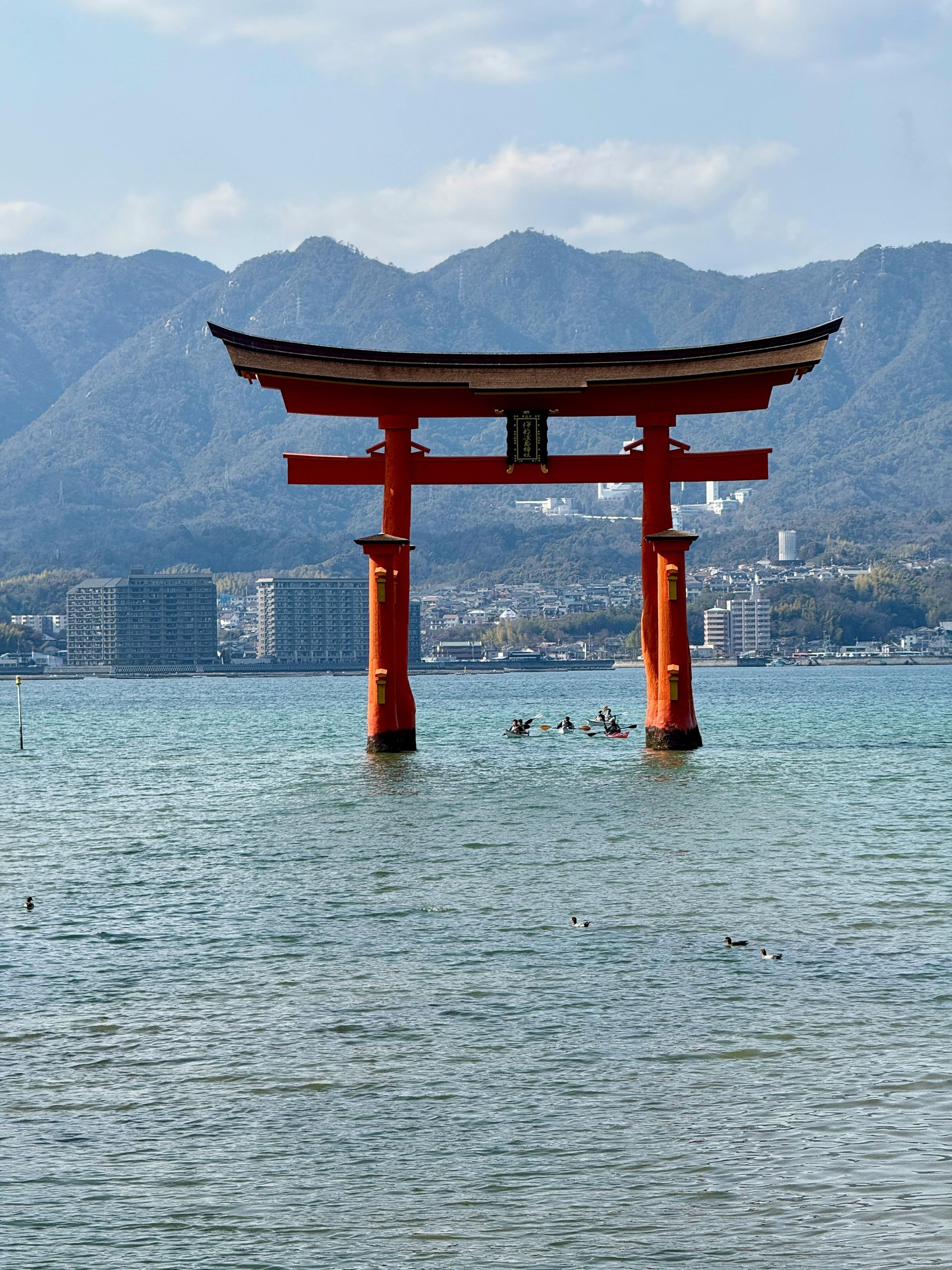 De iconische rode Itsukushima Torii-poort staat in het water, omringd door bergen onder een zonnige blauwe hemel.