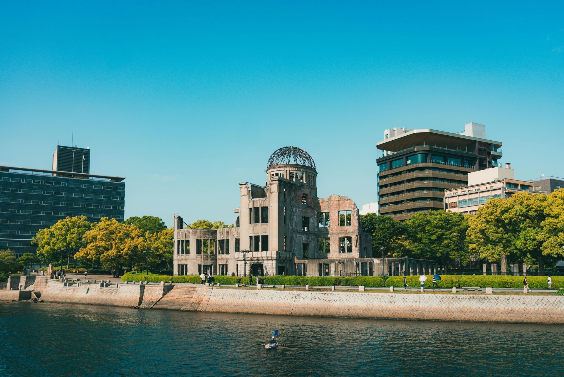 Het Vredesmonument van Hiroshima, een ruïne van een koepelvormig gebouw, staat op een met gras begroeide rivieroever onder een helderblauwe hemel.