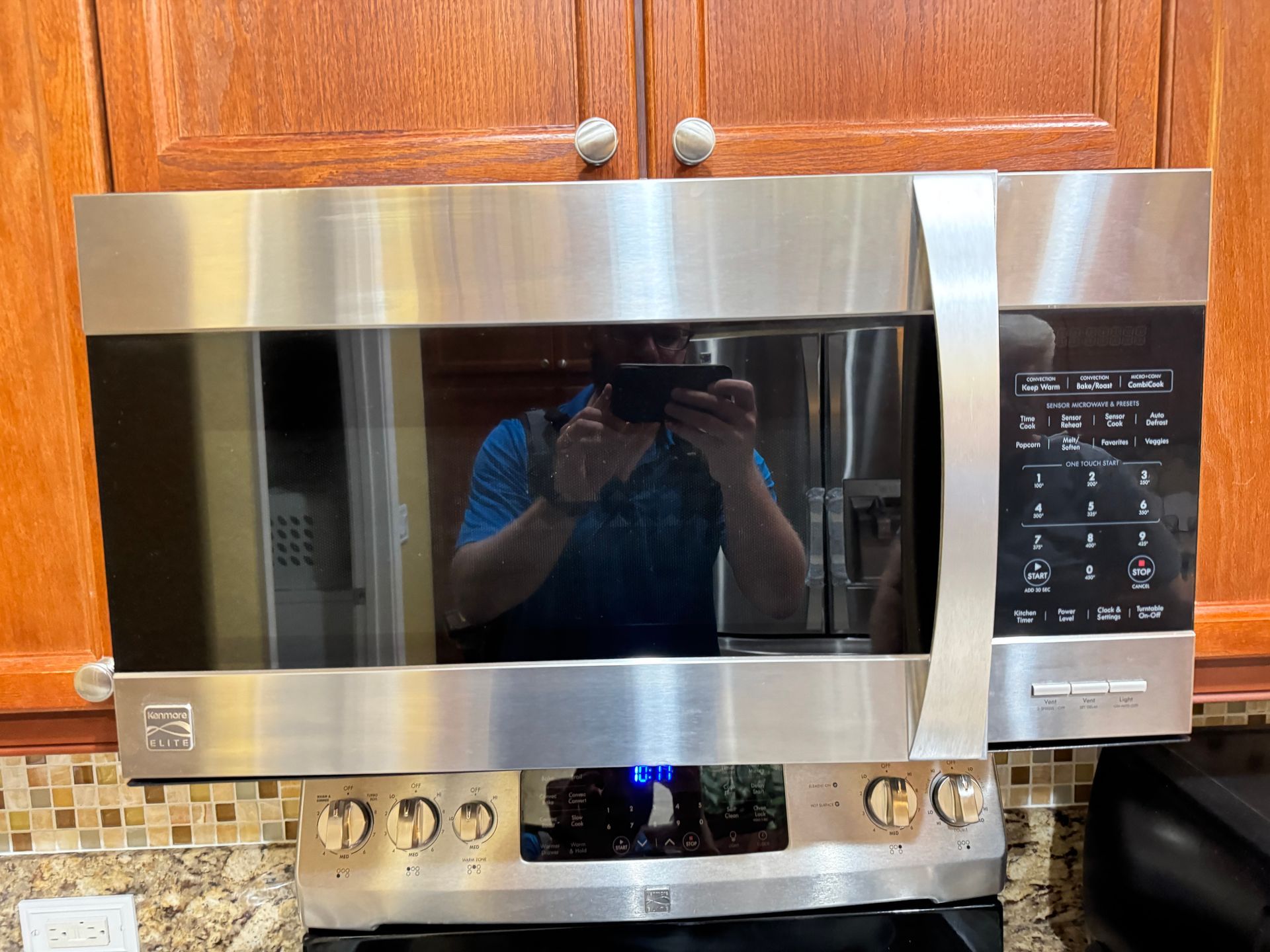 Stainless steel microwave above a stove in a kitchen with wooden cabinets and tiled backsplash.