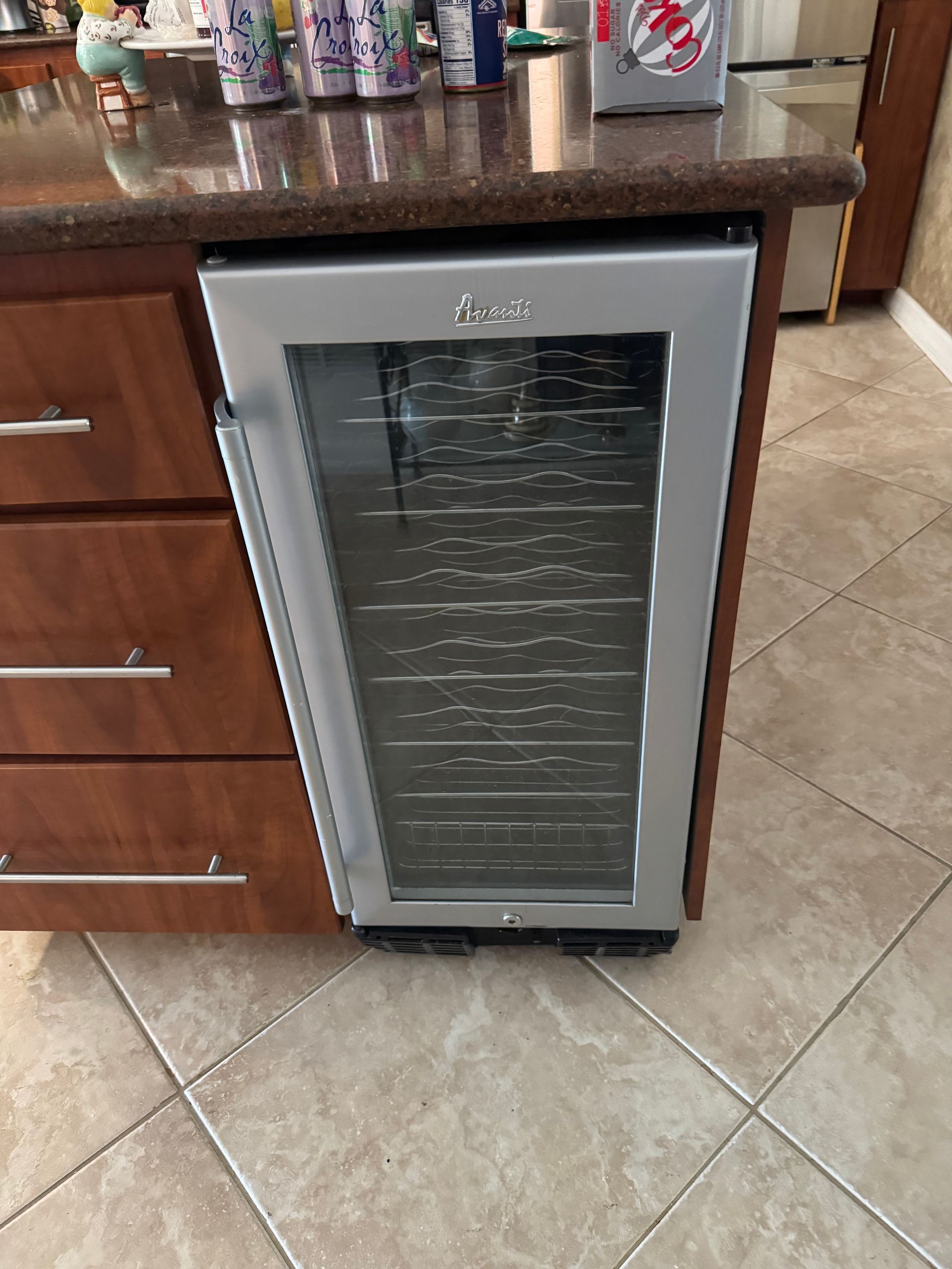 Small glass-front mini fridge under a kitchen counter on a tiled floor.