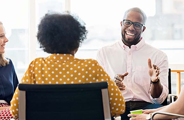 Group in a meeting room, smiling man gesturing while others listen around a table