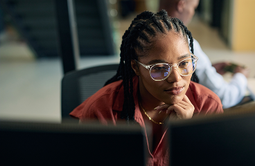 Person with glasses working at a computer in an office, resting chin on hand and looking focused