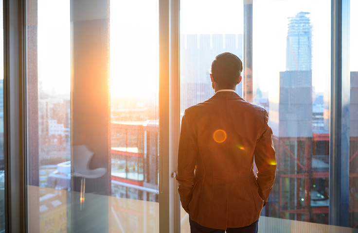 Person standing by a window overlooking a city skyline at sunset