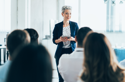 Presenter speaking to seated audience in a bright conference room, holding notes