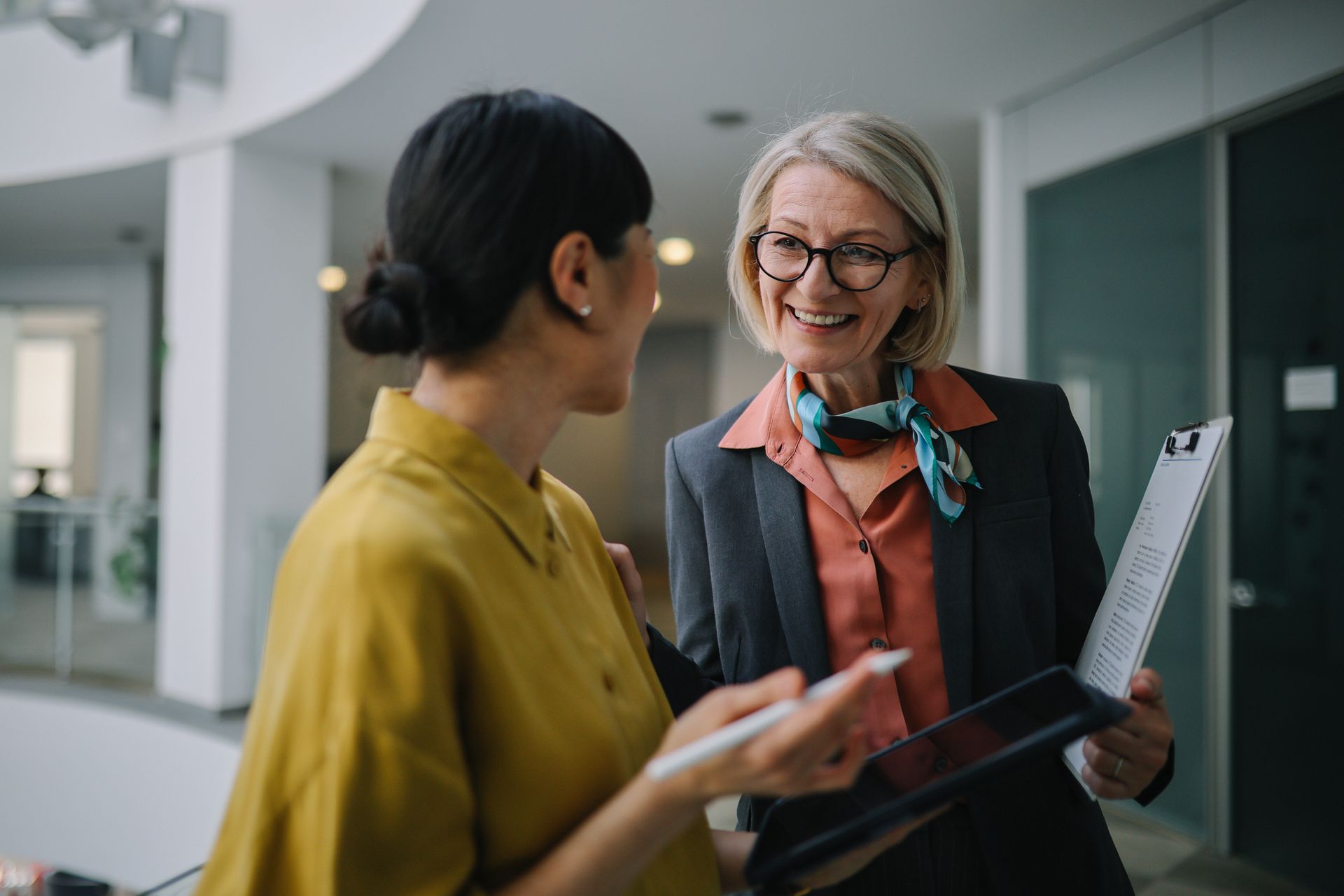 Two coworkers talking in an office, one holding a tablet and the other a clipboard, both smiling