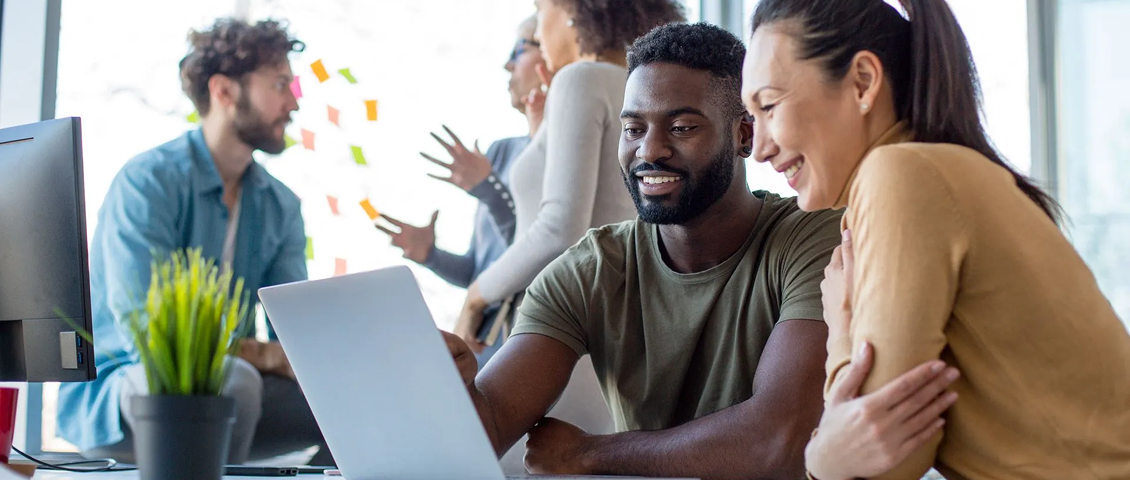 Three coworkers collaborating around a laptop in a bright office, one gesturing while others smile
