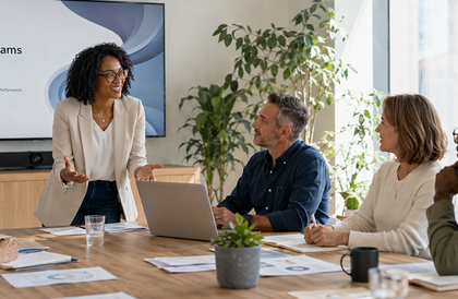 Three colleagues in a meeting room, one standing and presenting beside a laptop and papers on the table.