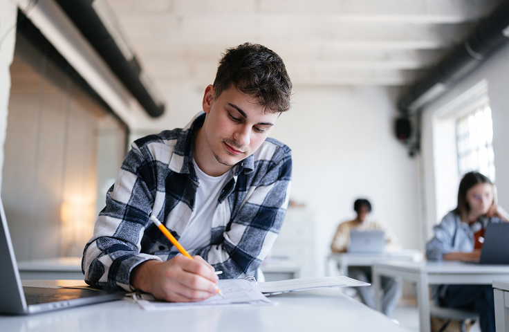 Student writing with a pencil at a desk in a bright classroom, with another student in the background