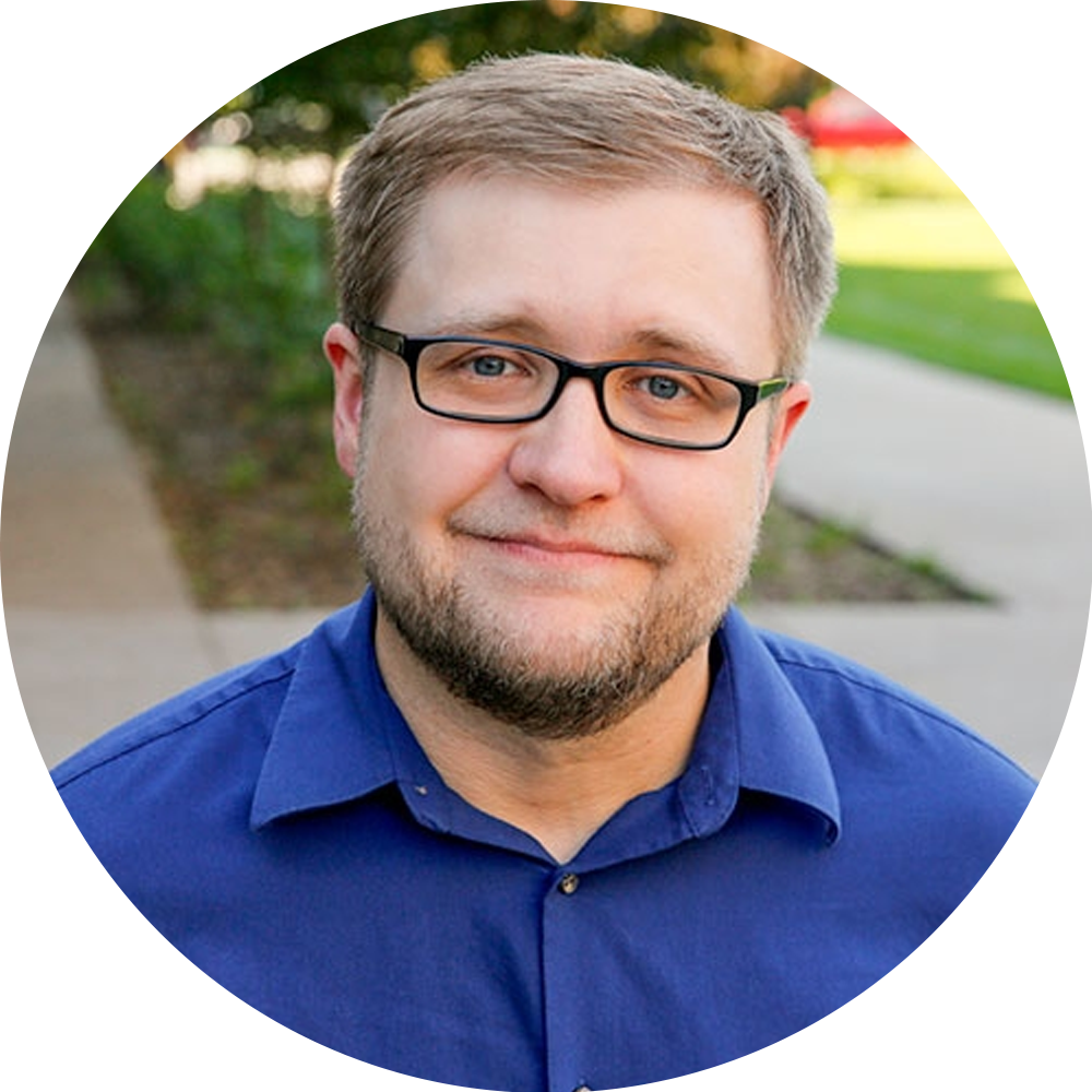 Man in glasses and blue shirt smiling outdoors on a sidewalk