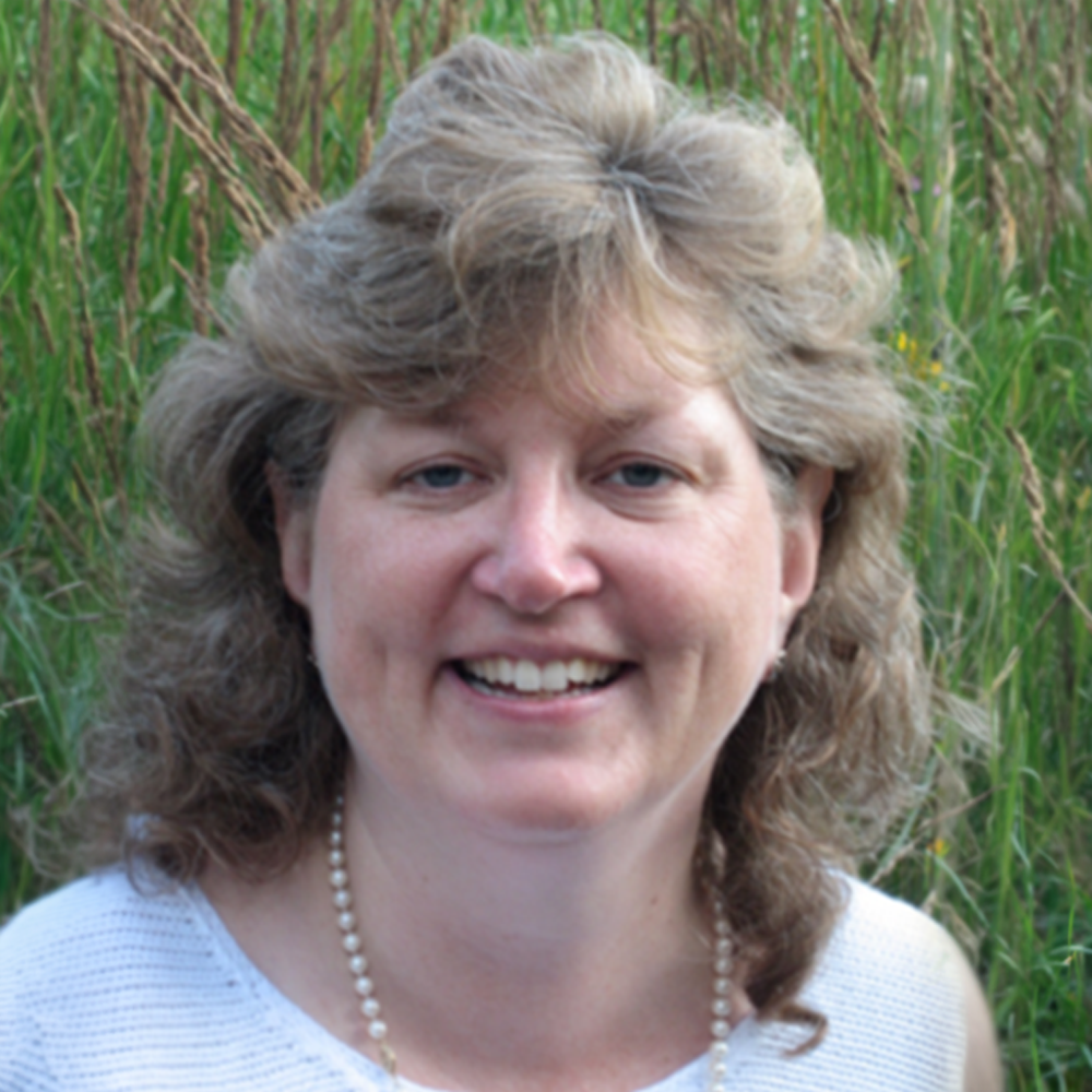 Smiling woman outdoors with wavy hair and a pearl necklace, standing in front of greenery