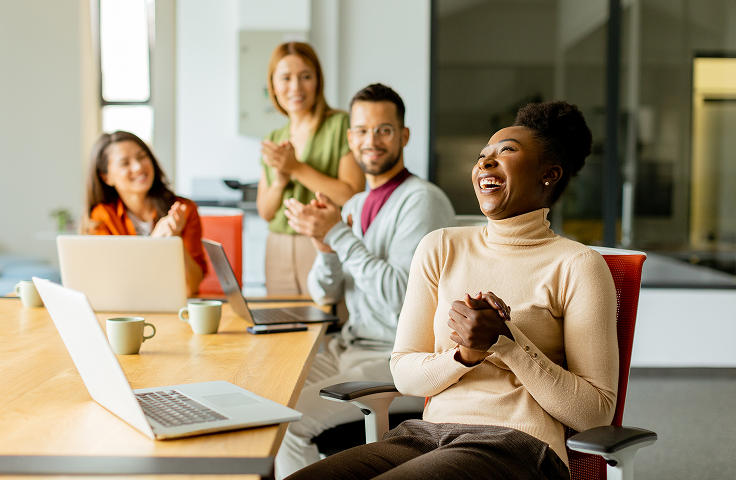 People in a bright office smiling and chatting around a conference table with laptops and coffee mugs