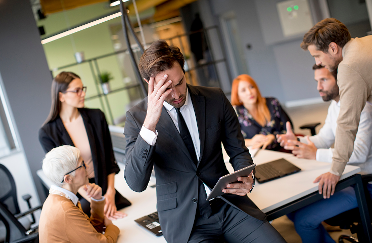 Business meeting with one man stressed at a table while coworkers discuss around him in an office