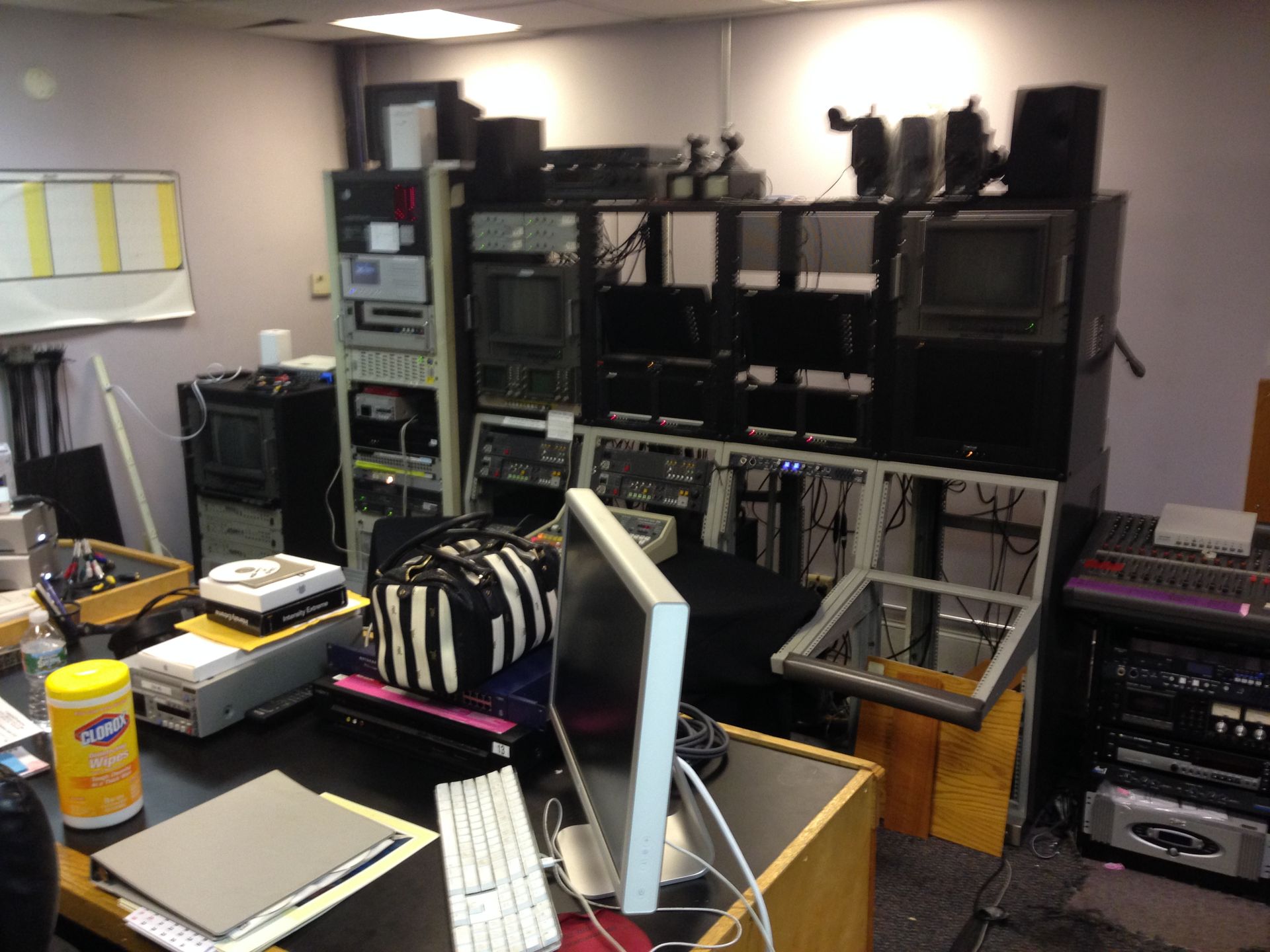 A cluttered control room with equipment racks, monitors, and a computer on a desk.