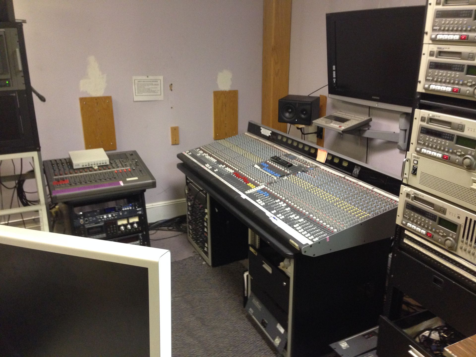 Recording studio with audio mixing console and rack-mounted equipment. Beige walls and monitors.