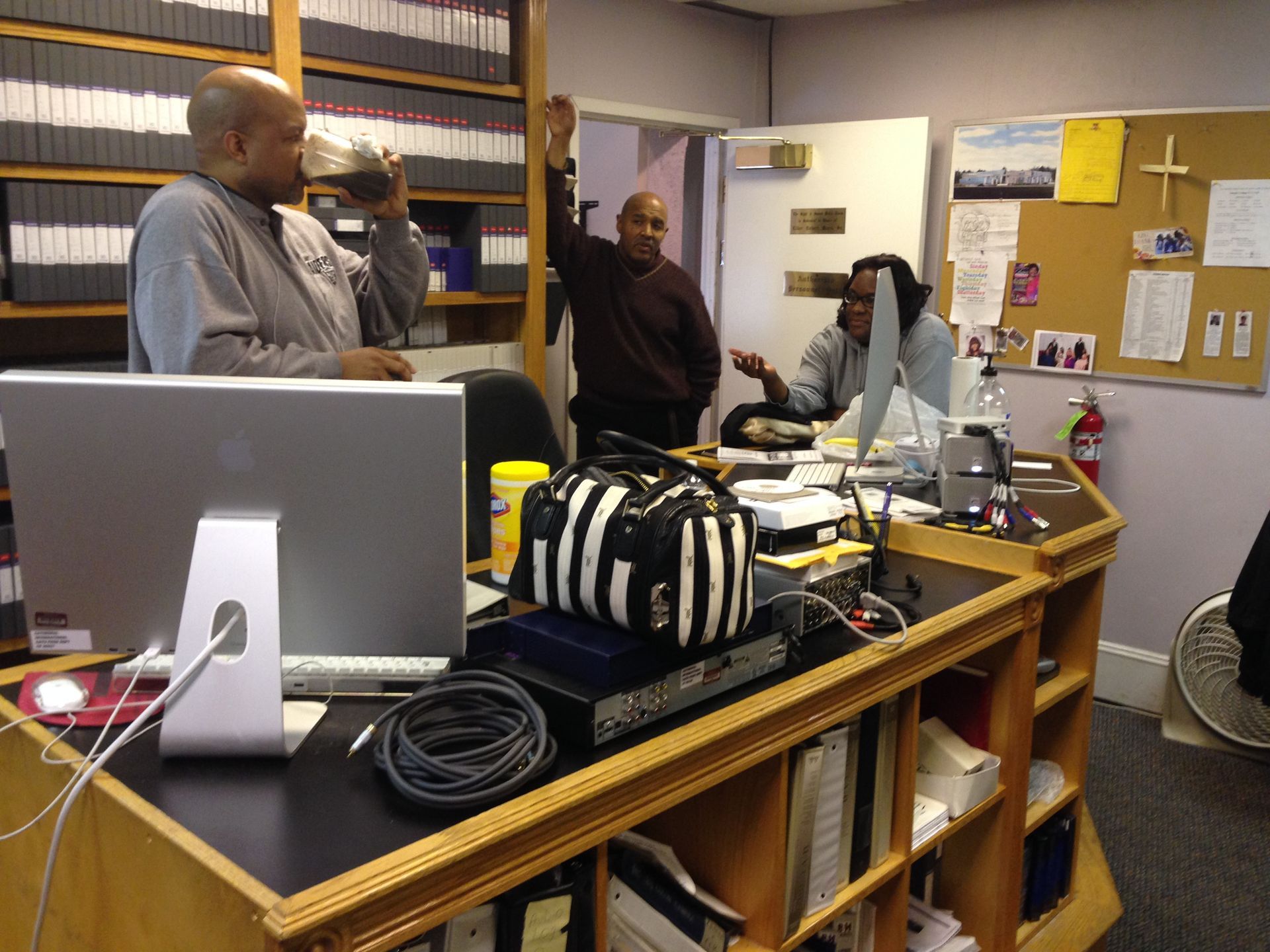 Three people in a cluttered office; one raises hand, others talk. Shelves of files in the background, computer visible.
