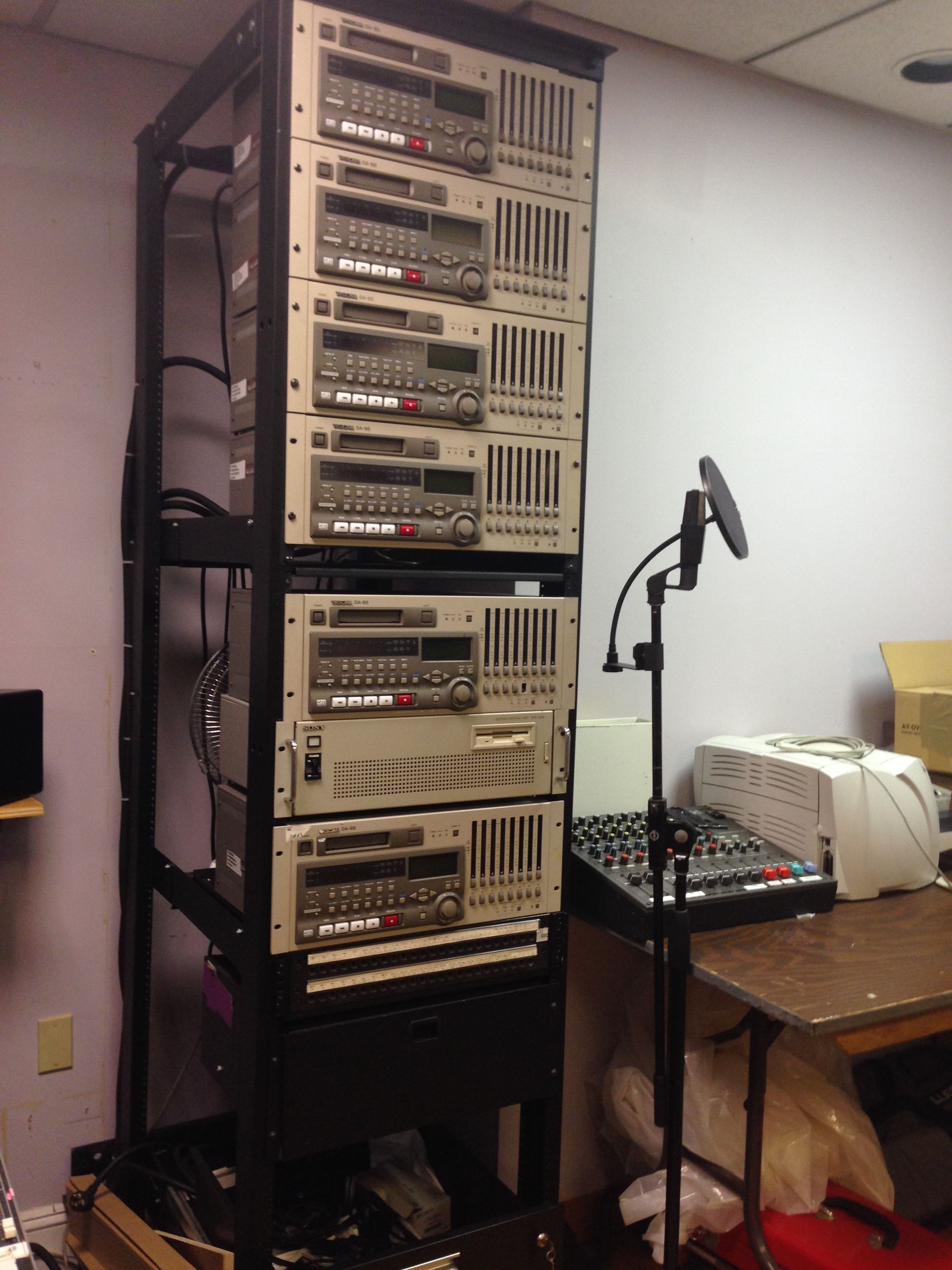 Black rack holding audio equipment; microphone on stand in front, small desk with mixing board.