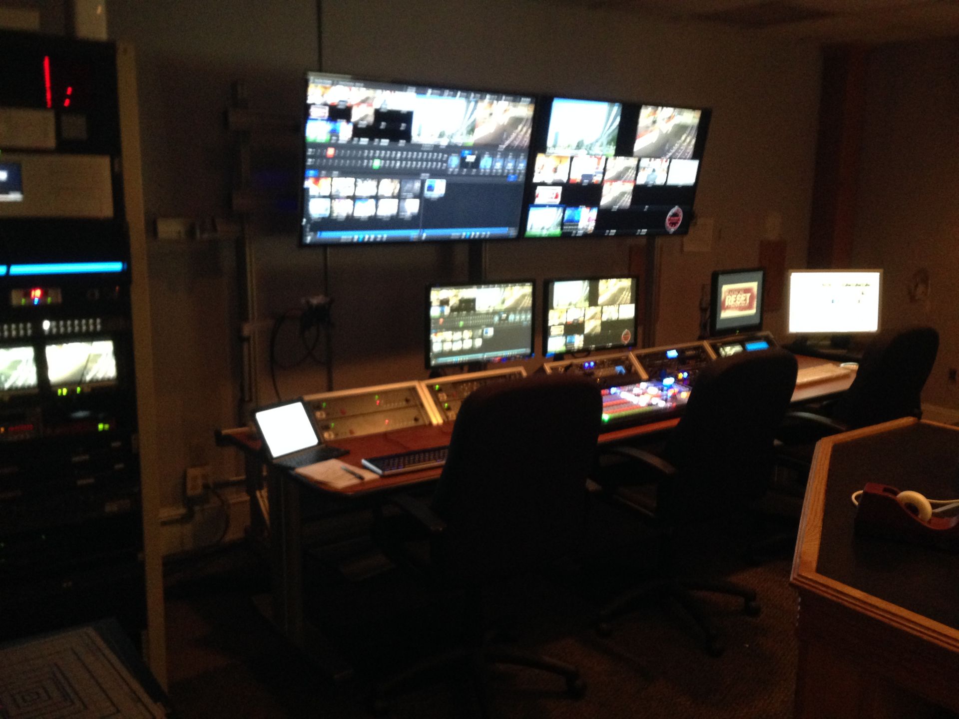 Control room with multiple screens, consoles, and chairs. Dark setting with technological equipment.
