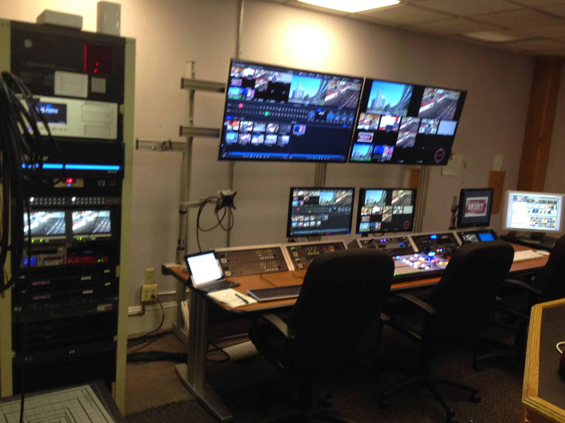 Control room with multiple monitors, equipment racks, and a long desk with chairs.