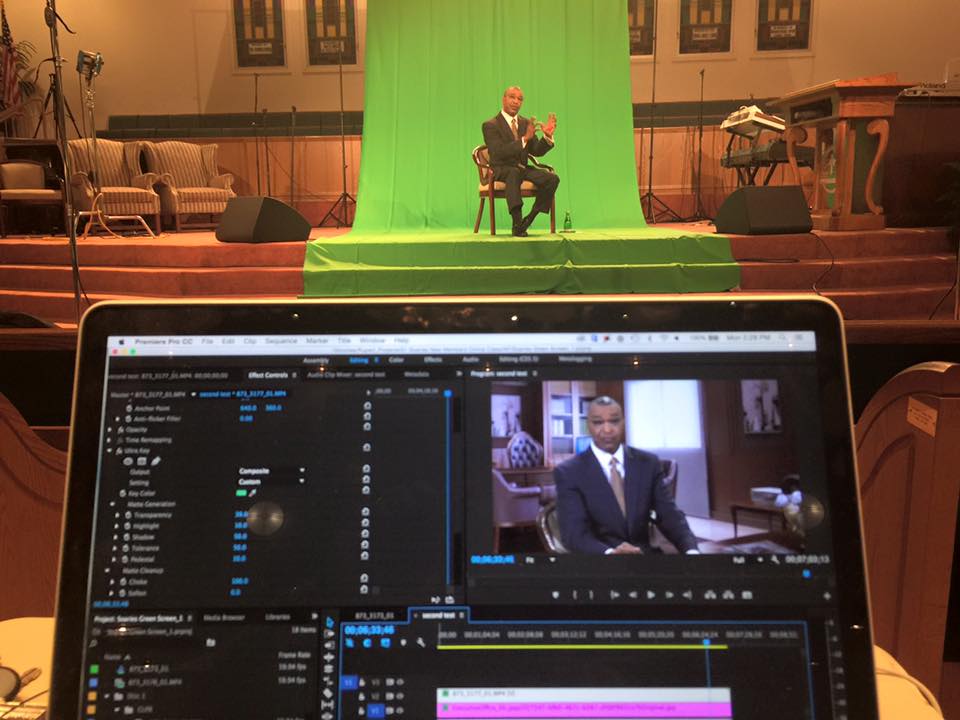 A man sits in front of a green screen, filming in a church. A laptop displays the edited footage.