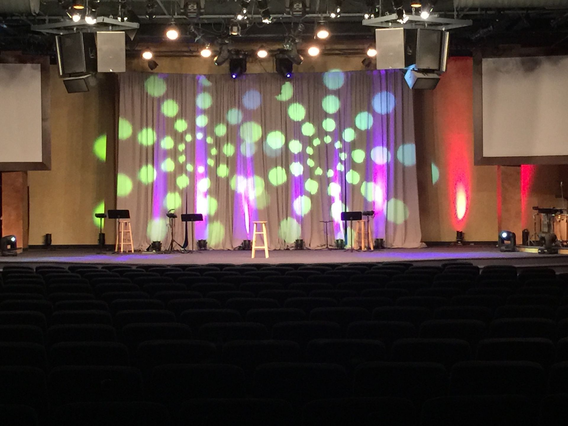 Stage with curtains and colorful spotlight projections. Empty chairs face the stage.
