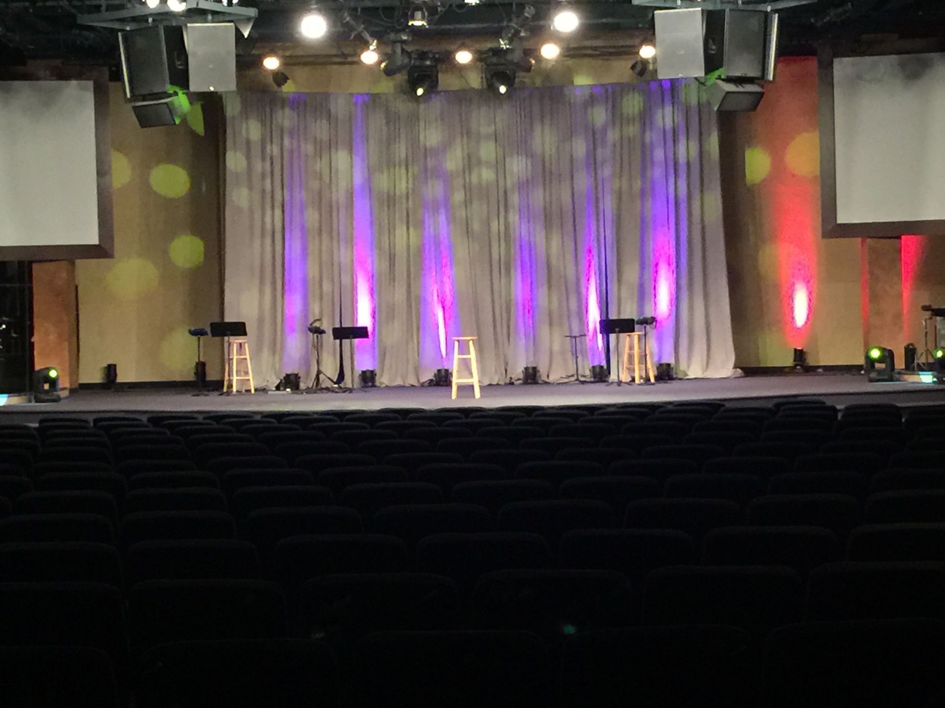 Stage with curtains lit with purple and red lights, a row of chairs in front.
