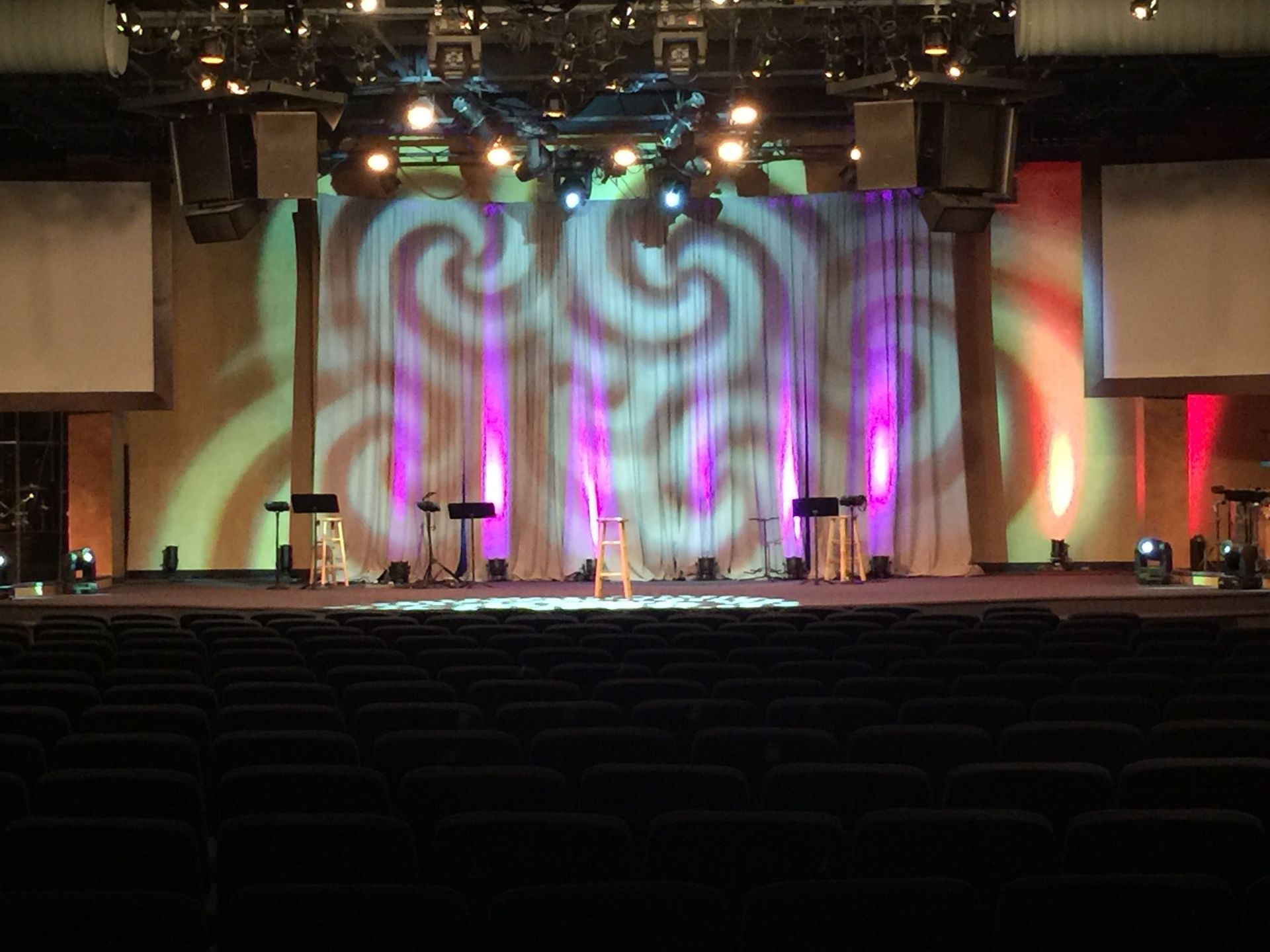 Auditorium stage with swirling light projections and microphones, viewed from the audience.