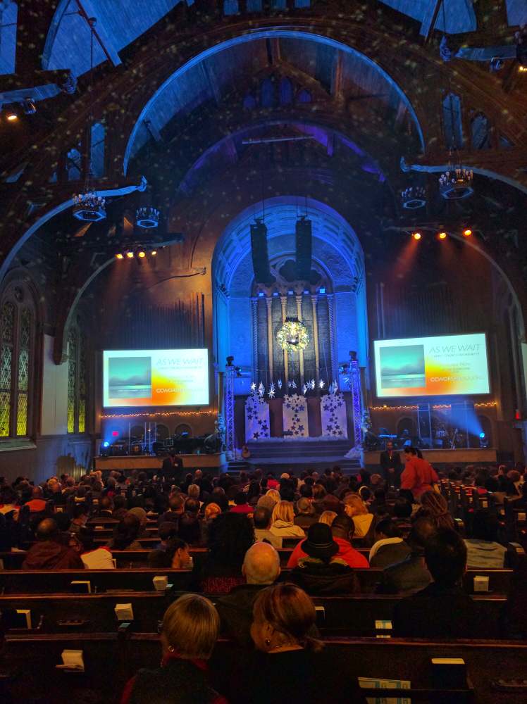 Large audience seated in a church; stage lit with blue hues and two screens displaying a landscape.