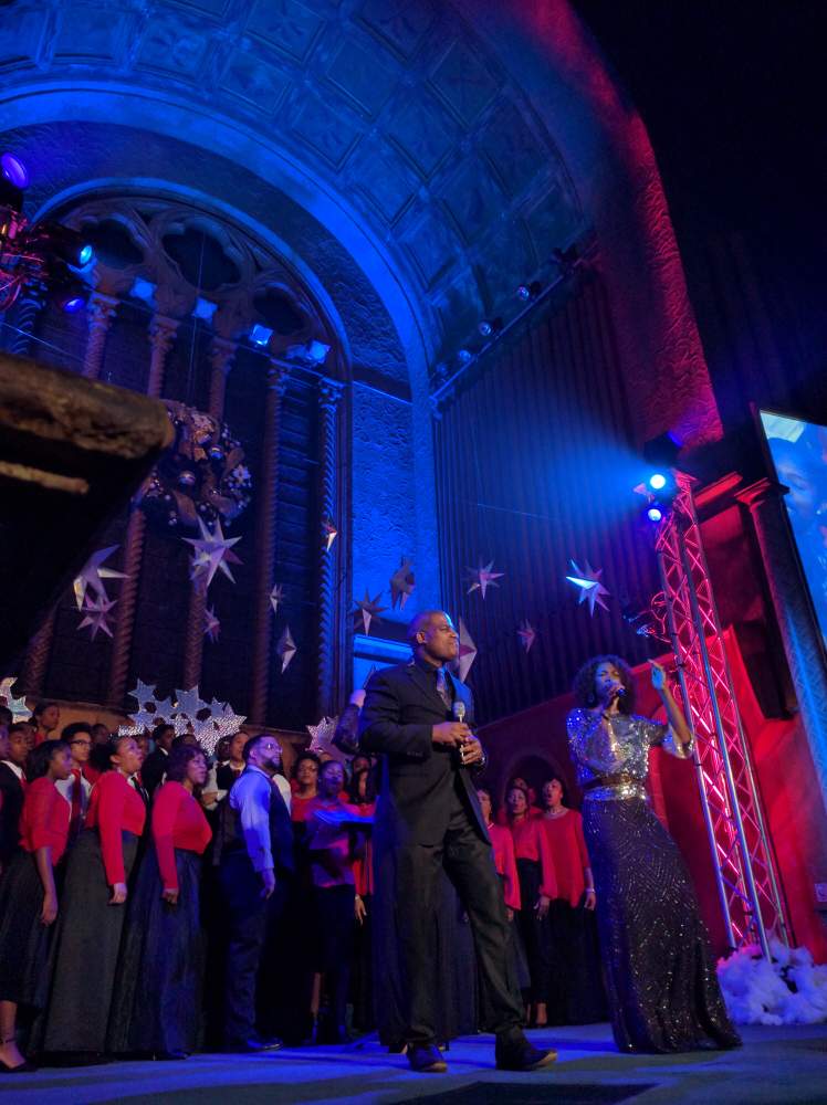Choir performance with two soloists on stage. Large ornate hall, blue and red lighting.