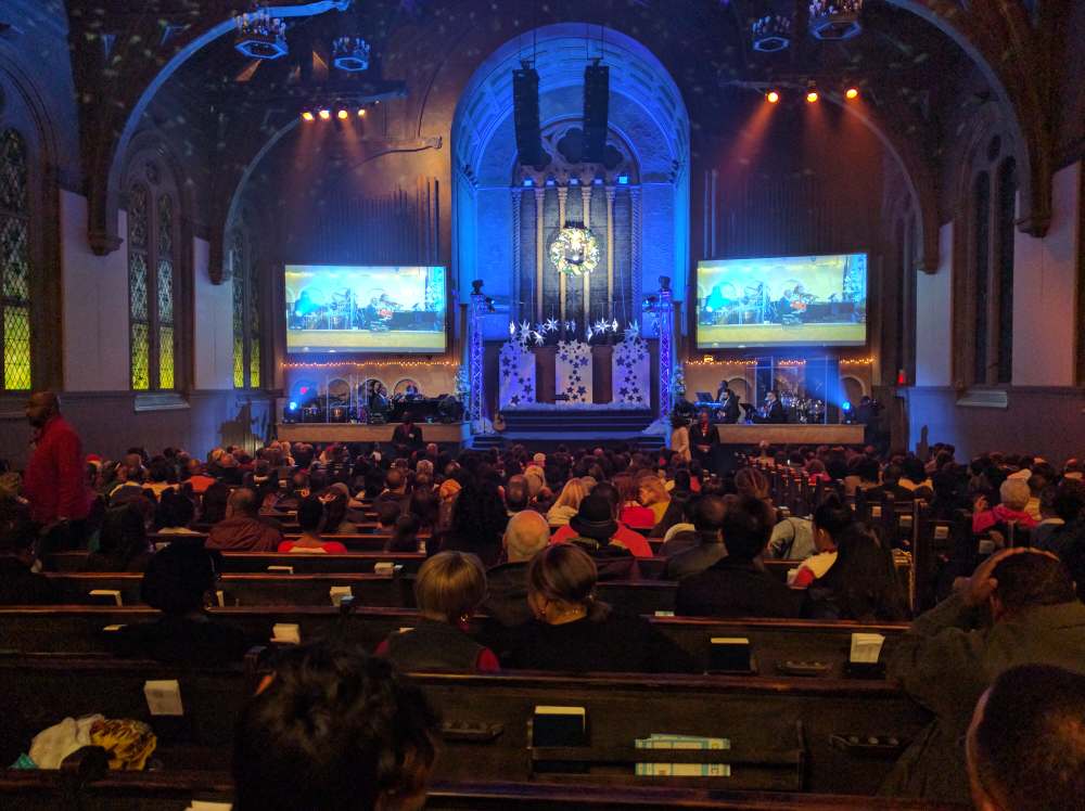 Audience in a dimly lit church watches a performance on a stage with screens and blue lighting.