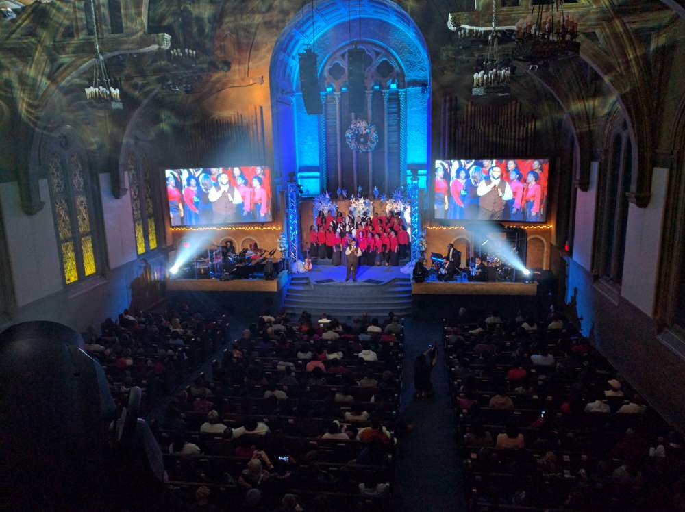 Choir performing on stage in a large hall with screens, audience watching. Blue lighting highlights the stage.