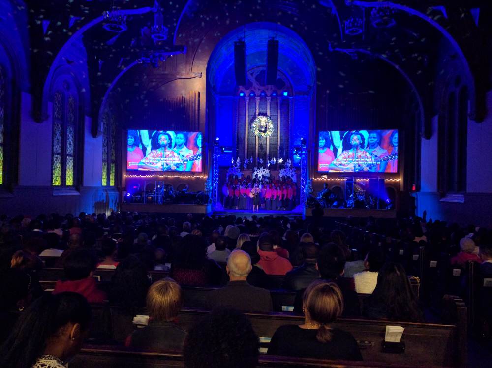 Audience in a church watches a stage with screens. Blue stage lights illuminate performers and the audience.